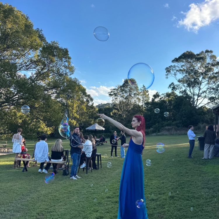 performer with red hair in blue dress making bubbles in a green grassy field with blue sky. You can see some bubbles formed around the bubble sticks and rope